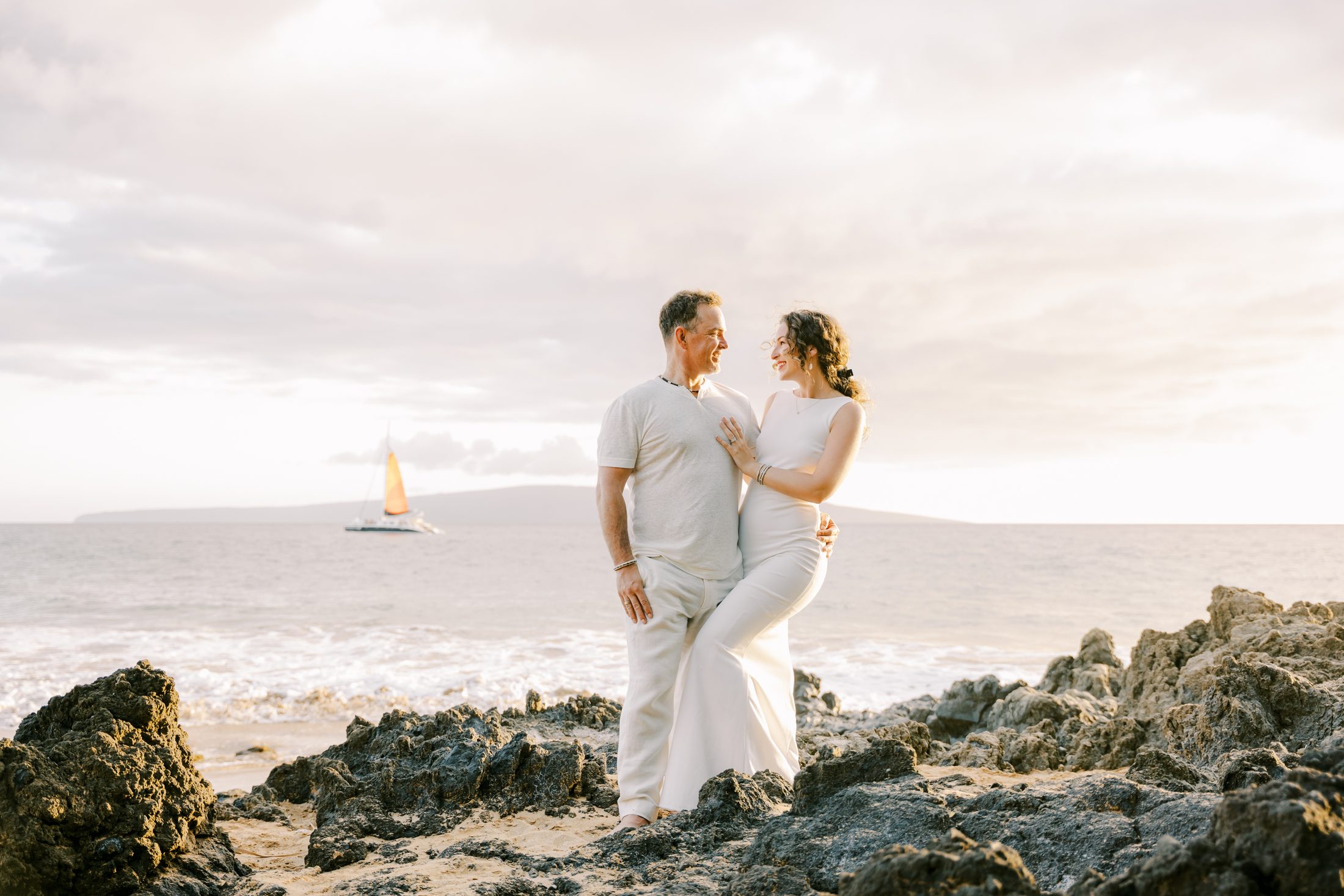 Pete and Danielle standing together on lava rocks at a Maui beach, a sailboat on the water behind them